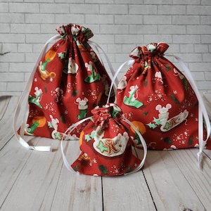 May include: Three red fabric drawstring bags with white ribbon ties. The bags are decorated with a Christmas-themed pattern of mice and gingerbread men. The bags are arranged in order of size, with the largest bag at the top and the smallest bag at the bottom.