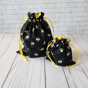May include: Two black fabric drawstring bags with yellow drawstrings. The bags are decorated with a repeating pattern of yellow and black bees. The larger bag is in the background and the smaller bag is in the foreground.