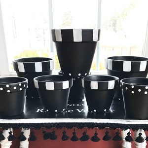 May include: A set of six black terracotta flower pots with white polka dot and striped patterns. The pots are arranged in a pyramid shape on a table with a black and white patterned tablecloth.