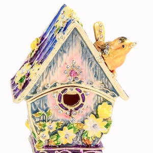 May include: A blue and pink birdhouse trinket box with a yellow bird perched on the roof. The birdhouse is decorated with flowers and rhinestones.