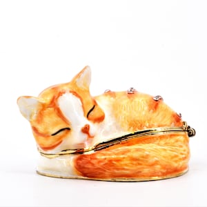 May include: A small, orange and white ceramic cat figurine with closed eyes, curled up in a sleeping position. The figurine has a gold trim and is decorated with rhinestones.