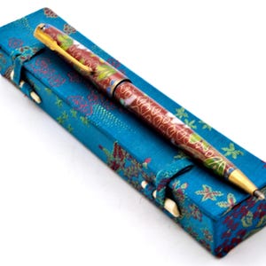 Puede incluir: Un bol&iacute;grafo decorativo con un dise&ntilde;o floral rojo y dorado descansa sobre un estuche azul. El bol&iacute;grafo tiene un clip y una punta dorados. El estuche es una caja rectangular con un estampado floral y un cierre.