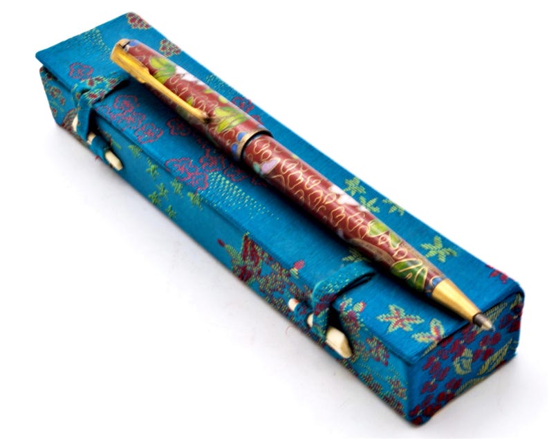 Puede incluir: Un bol&iacute;grafo decorativo con un dise&ntilde;o floral rojo y dorado descansa sobre un estuche azul. El bol&iacute;grafo tiene un clip y una punta dorados. El estuche es una caja rectangular con un estampado floral y un cierre.