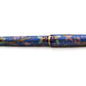 Puede incluir: Un bol&iacute;grafo azul con un dise&ntilde;o floral. El bol&iacute;grafo tiene una punta y detalles dorados. El patr&oacute;n floral incluye detalles en rosa, verde y dorado. El bol&iacute;grafo est&aacute; sobre un fondo blanco.