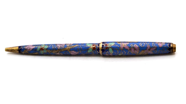 Puede incluir: Un bol&iacute;grafo azul con un dise&ntilde;o floral. El bol&iacute;grafo tiene una punta y detalles dorados. El patr&oacute;n floral incluye detalles en rosa, verde y dorado. El bol&iacute;grafo est&aacute; sobre un fondo blanco.
