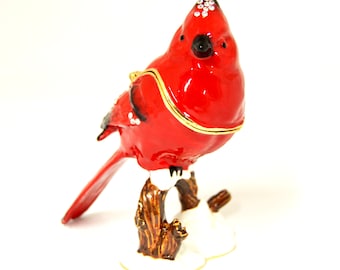 May include: A red cardinal bird figurine with a gold trim and rhinestones on its head. The bird is perched on a branch with snow on the ground. The figurine is made of metal and has a hinged lid, making it a trinket box.