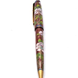 Puede incluir: Un bol&iacute;grafo decorativo con un cuerpo granate, adornado con motivos florales blancos y morados y hojas verdes. El bol&iacute;grafo presenta un clip y una punta dorados, y un acento azul cerca de la punta. Es un instrumento de escritura.