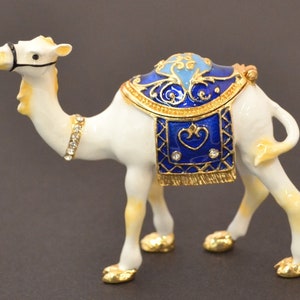 May include: A white camel figurine with a blue and gold saddle. The camel is decorated with rhinestones and has a gold trim.
