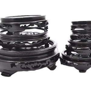 May include: Two stacks of black wooden display stands. The stands have a round shape with carved floral designs and are of varying sizes. The stands are made of a dark, glossy wood and are set against a white background.