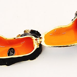 Puede incluir: Caja de joyer&iacute;a de teckel abierta en esmalte negro y dorado con interior naranja. La caja tiene un cierre magn&eacute;tico y un logotipo de Ciel en la parte inferior.