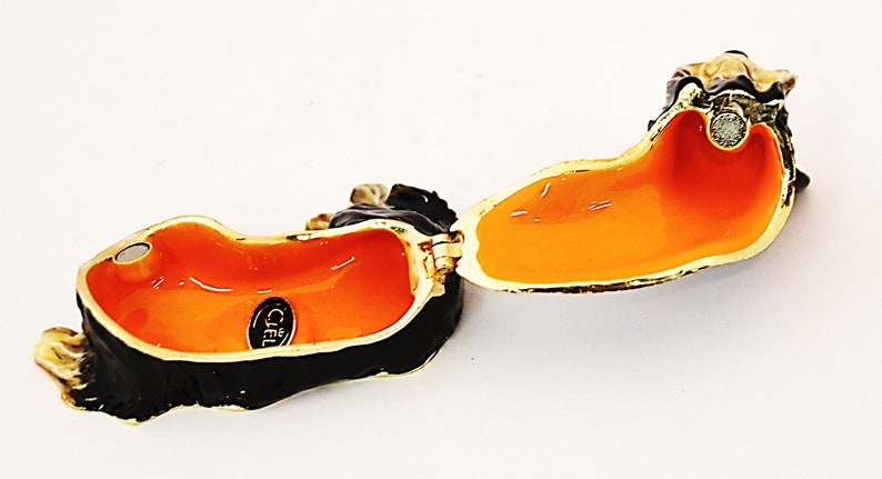Puede incluir: Caja de joyer&iacute;a de teckel abierta en esmalte negro y dorado con interior naranja. La caja tiene un cierre magn&eacute;tico y un logotipo de Ciel en la parte inferior.