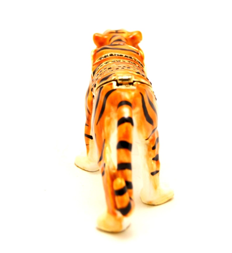 May include: A ceramic tiger figurine with orange and black stripes. The figurine is standing on a white surface and is facing away from the camera.