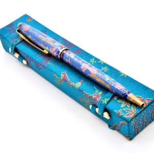 Puede incluir: Un bol&iacute;grafo azul con un dise&ntilde;o floral descansa sobre un estuche a juego. El bol&iacute;grafo tiene detalles dorados y un patr&oacute;n detallado en azul, rosa y dorado. El estuche es rectangular con un patr&oacute;n floral y cierres blancos.