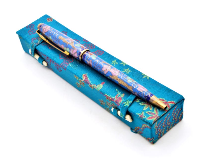Puede incluir: Un bol&iacute;grafo azul con un dise&ntilde;o floral descansa sobre un estuche a juego. El bol&iacute;grafo tiene detalles dorados y un patr&oacute;n detallado en azul, rosa y dorado. El estuche es rectangular con un patr&oacute;n floral y cierres blancos.