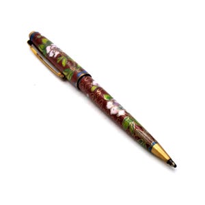 Puede incluir: Bol&iacute;grafo decorativo con cuerpo granate, adornado con un estampado floral en blanco, verde y azul. El bol&iacute;grafo presenta detalles dorados en el clip y la punta, y una punta de escritura negra. El bol&iacute;grafo est&aacute; sobre un fondo blanco.