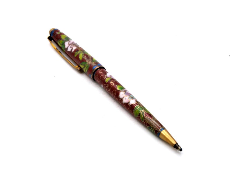 Puede incluir: Bol&iacute;grafo decorativo con cuerpo granate, adornado con un estampado floral en blanco, verde y azul. El bol&iacute;grafo presenta detalles dorados en el clip y la punta, y una punta de escritura negra. El bol&iacute;grafo est&aacute; sobre un fondo blanco.