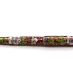 Puede incluir: Bol&iacute;grafo decorativo con dise&ntilde;o floral. El bol&iacute;grafo presenta un fondo granate con flores blancas y moradas, hojas verdes y detalles dorados. La punta y el extremo del bol&iacute;grafo son dorados.