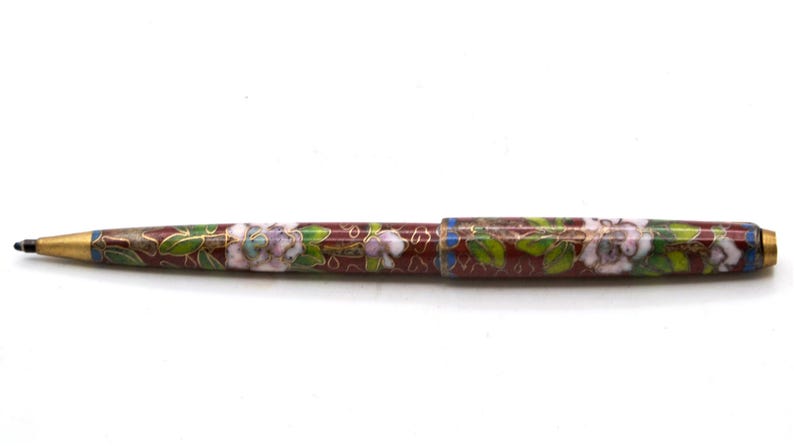Puede incluir: Bol&iacute;grafo decorativo con dise&ntilde;o floral. El bol&iacute;grafo presenta un fondo granate con flores blancas y moradas, hojas verdes y detalles dorados. La punta y el extremo del bol&iacute;grafo son dorados.