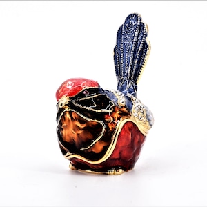 May include: A small, intricately detailed bird figurine made of metal. The bird is painted in shades of red, black, gold, and blue. The bird's tail feathers are spread out and have a blue and gold design. The bird's head is tilted to the side, and its eyes are made of small, clear stones.