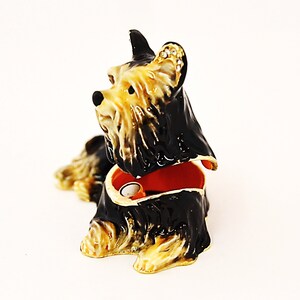 Puede incluir: Una figurita de Yorkshire Terrier negra y marr&oacute;n con una tapa con bisagras, que revela un peque&ntilde;o espacio interior. La figurita est&aacute; hecha de metal y tiene un dise&ntilde;o detallado.