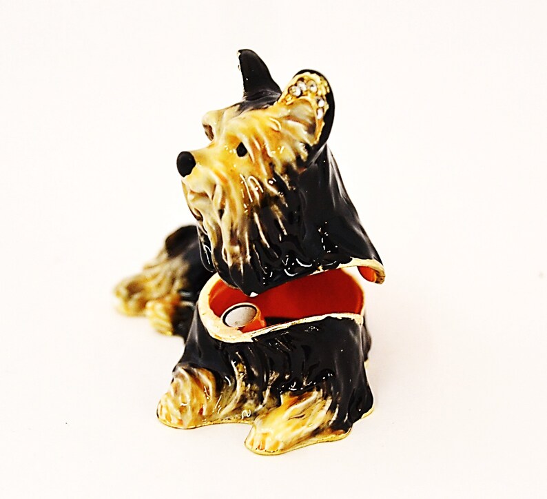 Puede incluir: Una figurita de Yorkshire Terrier negra y marr&oacute;n con una tapa con bisagras, que revela un peque&ntilde;o espacio interior. La figurita est&aacute; hecha de metal y tiene un dise&ntilde;o detallado.