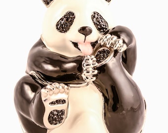Puede incluir: Una figurita de panda blanco y negro con una expresión juguetona, sentada sobre sus ancas y sosteniendo su pata en su boca. La figurita está hecha de metal y tiene un acabado brillante, similar al esmalte. Los ojos del panda son negros y brillantes, y su lengua sobresale.