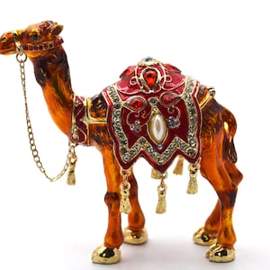 May include: A golden camel figurine with a red and gold jeweled saddle. The camel is standing on a white background.