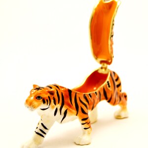 May include: A small, orange and black tiger figurine with a hinged back that opens to reveal a small compartment. The figurine is made of porcelain and has a glossy finish.