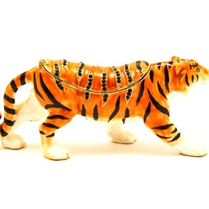 May include: A detailed figurine of a tiger, with orange and black stripes, standing on a white surface. The tiger is made of metal and has a jeweled design on its back.