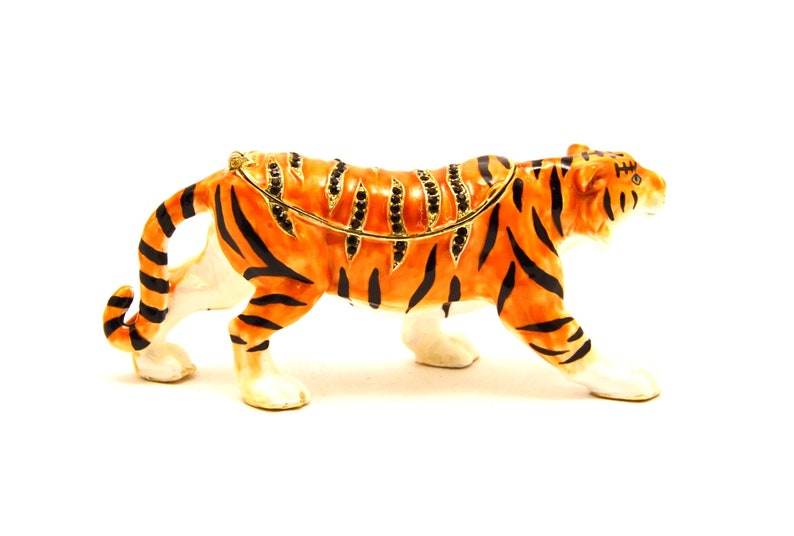 May include: A detailed figurine of a tiger, with orange and black stripes, standing on a white surface. The tiger is made of metal and has a jeweled design on its back.