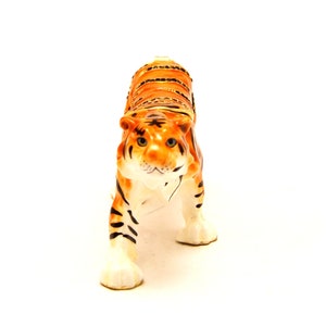 May include: A ceramic tiger figurine with orange and black stripes. The tiger has a white face with black eyes and a pink nose.