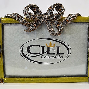 Puede incluir: Un marco de fotos verde con un lazo adornado con joyas y el texto "CIEL Collectables" en la parte delantera.