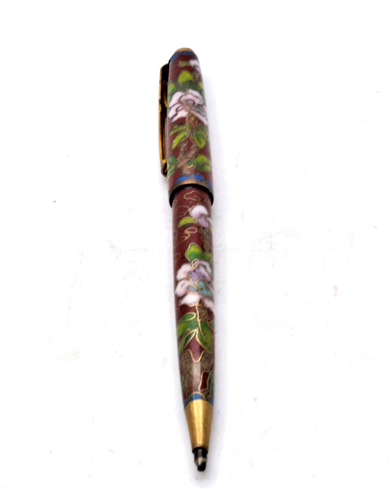 Puede incluir: Un bol&iacute;grafo decorativo con un dise&ntilde;o floral. El bol&iacute;grafo presenta un color base rojo oscuro con patrones florales blancos, verdes y azules. El bol&iacute;grafo tiene un clip y una punta dorados.