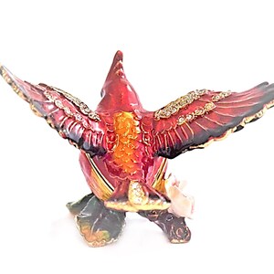 May include: A red cardinal bird with a jeweled design and outstretched wings, holding a pink rose in its beak. The bird is perched on a green stem with leaves. The figurine is made of metal and has a detailed, intricate design.