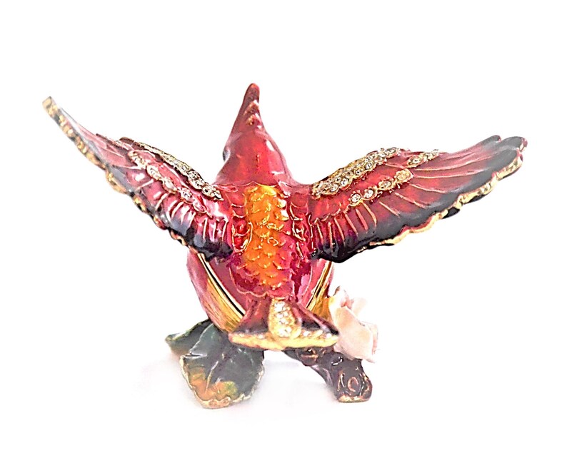 May include: A red cardinal bird with a jeweled design and outstretched wings, holding a pink rose in its beak. The bird is perched on a green stem with leaves. The figurine is made of metal and has a detailed, intricate design.