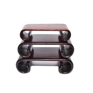 May include: A three-tiered, dark wood display stand with a curved, scroll-like design. The stand is a rich, reddish-brown colour, with each tier supported by elegantly curved legs. The design is simple yet sophisticated.