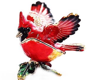 May include: A red cardinal figurine with outstretched wings, perched on a green holly leaf. The figurine is made of metal and has a detailed design.