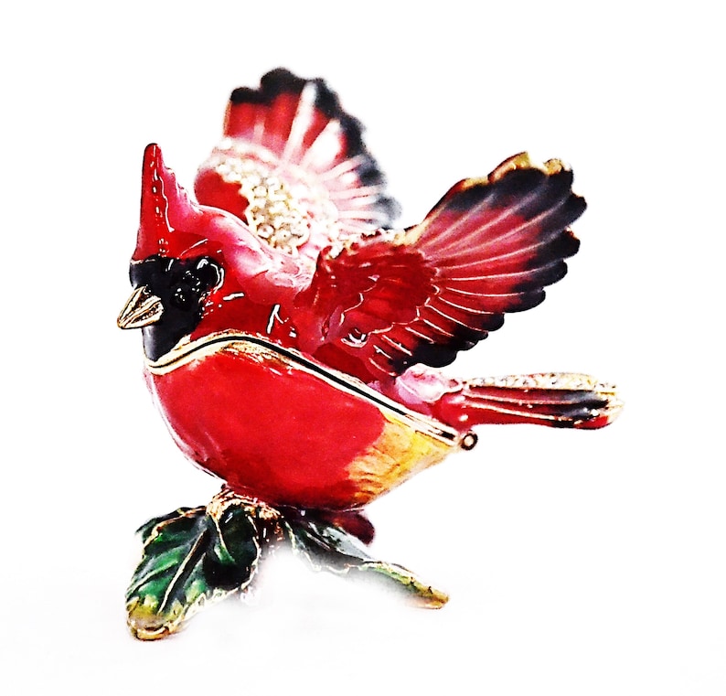 May include: A red cardinal figurine with outstretched wings, perched on a green holly leaf. The figurine is made of metal and has a detailed design.
