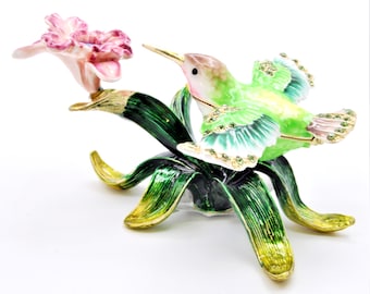 May include: A green and pink hummingbird with jeweled accents perched on a green and gold flower with pink petals.