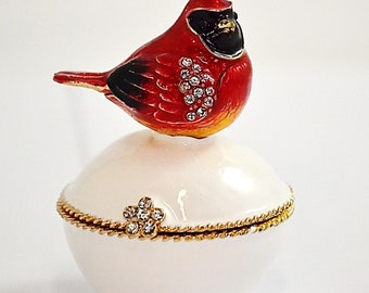 May include: A white ceramic trinket box with a red cardinal bird figurine on top. The cardinal has a black head and a gold beak. The box has a gold trim and a small flower design with rhinestones.