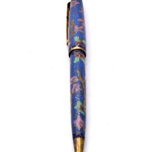 Puede incluir: Un bol&iacute;grafo azul con un dise&ntilde;o floral. El bol&iacute;grafo presenta un clip y una punta dorados, y el cuerpo est&aacute; adornado con un patr&oacute;n de flores y hojas rosas, verdes y doradas. El bol&iacute;grafo est&aacute; sobre un fondo blanco.