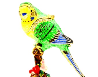 May include: A green and yellow parakeet figurine with blue tail feathers perched on a brown branch with red flowers. The figurine is made of metal and has a shiny finish.