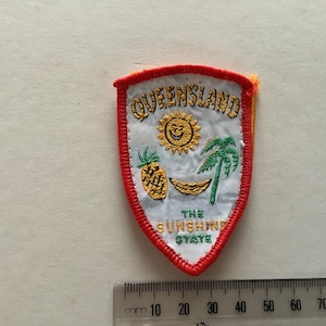 Op de afbeelding: Geborduurde patch in schildvorm met het woord "QUEENSLAND" boven een lachende zon, ananas, banaan en palmboom. De tekst "THE SUNSHINE STATE" staat eronder. De patch heeft een rode rand.