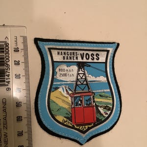 May include: A shield-shaped patch with a blue border and a black backing. The patch features a red cable car, mountains, and the text "HANGURS-BANEN VOSS". It also includes the text "800 m.o.h. 2500 f.o.h."