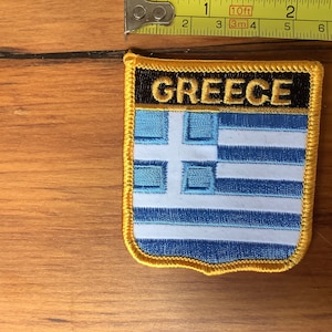 Puede incluir: Un parche bordado amarillo con la bandera griega y la palabra "GREECE" en letras negras.