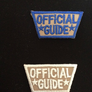 Puede incluir: Dos parches bordados con el texto "OFFICIAL GUIDE" en color tostado con costuras azules y blancas. El parche superior tiene un borde azul y el parche inferior tiene un borde blanco.