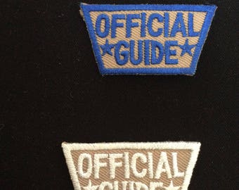 Puede incluir: Dos parches bordados con el texto "OFFICIAL GUIDE" en color tostado con costuras azules y blancas. El parche superior tiene un borde azul y el parche inferior tiene un borde blanco.
