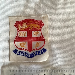 May include: A white fabric patch with a red shield featuring a gold lion, a red cross, and gold palm trees. The shield is surrounded by a blue border with the text "SUVA FIJI" in white.
