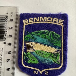 Puede incluir: Parche de fieltro azul con borde dorado y la palabra "BENMORE" en la parte superior. El parche presenta un paisaje con una presa, montañas y agua. Las letras "NZ" están en la parte inferior.