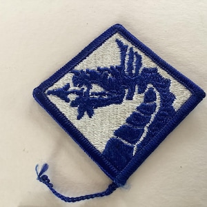 Può includere: Toppa ricamata a forma di diamante con un disegno di drago blu su sfondo bianco, incorniciata da un bordo blu. La toppa presenta un'illustrazione dettagliata di un drago. Un filo blu si estende dall'angolo inferiore.